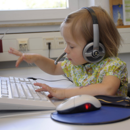 Aufnahme eines blonden Kindes mit einem Logitech-Headset auf dem Kopf, welches an einer weißen Tastatur sitzt. Das Kind ist mitten in der Bewegung des Tippens und wirkt sehr konzentriert.